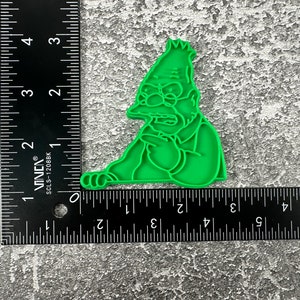 May include: Green plastic cookie cutter shaped like a cartoon character with a grumpy expression. The character is wearing a suit and has a large nose and glasses.