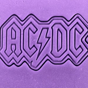 May include: A purple background with a black outline of the AC/DC logo. The logo is in a bold, blocky font with a lightning bolt between the C and the D.