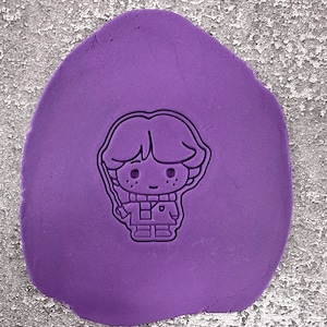 Könnte beinhalten: Ein lilafarbener Polymer Clay mit einer schwarzen Umrandung eines Comic-Charakters. Der Charakter ist ein Junge mit kurzen Haaren, der einen Schal und eine Jacke trägt.