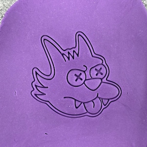 May include: A purple clay imprint of a cartoon cat with X's for eyes and a tongue sticking out.