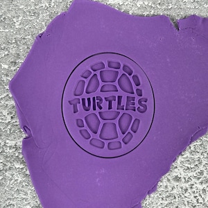May include: Purple play dough with a round cookie cutter pressed into it. The cookie cutter is a turtle shell with the word "TURTLES" in the center.