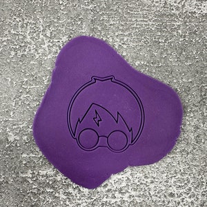 Könnte beinhalten: Lilafarbener Fondant mit einer schwarzen Umrandung eines Charakters, der runde Brillen und einen Blitz auf der Stirn trägt.