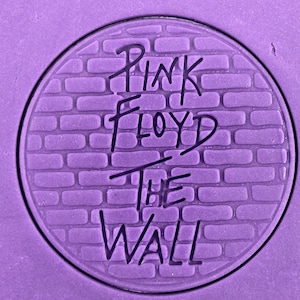 Könnte beinhalten: Ein lilafarbener Hintergrund mit einem schwarzen und weißen Kreis in der Mitte. Der Kreis hat ein Ziegelmuster und die Worte "Pink Floyd The Wall" sind in Schwarz geschrieben.