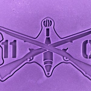 May include: A purple rubber mold with a design of two crossed rifles, a mortar shell, and the text "11C".