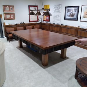 May include: A large, rectangular wooden pool table with a dark brown finish. The table has four sturdy, square legs and is covered with a black fabric skirt. The room features a light-colored carpet, wooden cabinets, and framed artwork.