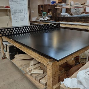 May include: A large, black ping pong table with a perforated net. The table is set on a wooden frame with a shelf underneath. The workshop setting includes various tools and materials.