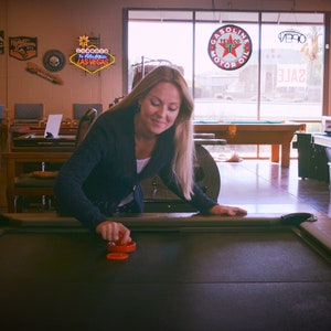 Może przedstawiać: Kobieta grająca w hokeja na powietrzu na czarnym stole do hokeja na powietrzu. Ma na sobie niebieski sweter i długie blond włosy. Stół ma na sobie dwa czerwone krążki.