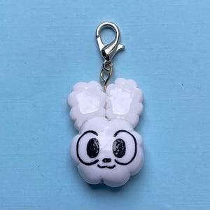 Op de afbeelding: Witte hanger met een cartoon gezicht en konijnenoren. De hanger heeft een zilveren clip aan de bovenkant.