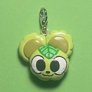 Op de afbeelding: Een lichtgroene bedel met een cartoon-stijl gezicht, met grote witgerande ogen en een kleine neus. De bedel heeft een zilveren sluiting en een groene achtergrond. Het ontwerp van de bedel omvat lichtbruine oorachtige vormen en een groen blad detail.