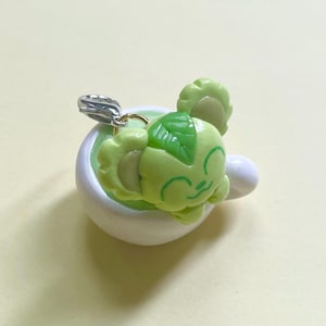 May include: A light green charm of a smiling cartoon character with a leaf on its head, resting in a white teacup. The charm has a silver clasp. The teacup is filled with a light green liquid.