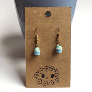 Puede incluir: Un par de pendientes colgantes de oro con un dije de cupcake azul y azul claro. Los pendientes se muestran en una tarjeta marrón con una nube de dibujos animados.