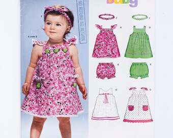 May include: A sewing pattern for baby clothes, featuring a pink floral dress, green gingham dress, white dress with a bow, and a polka dot dress. The pattern also includes instructions for making matching bloomers.