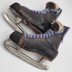 May include: A pair of vintage ice skates with brown leather boots and silver blades. The boots have blue and white striped laces and a worn appearance. The interior of the boots is lined with a light-colored, fuzzy material.
