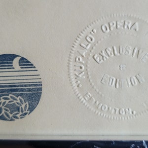 May include: Close-up of a framed print with two circular designs. The left circle features a blue design of a crescent moon, horizontal lines, and a wreath. The right circle reads "KUPALO OPERA EXCLUSIVE EDITION PREMIERE EDMONTON".