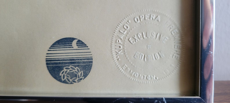 May include: Close-up of a framed print with two circular designs. The left circle features a blue design of a crescent moon, horizontal lines, and a wreath. The right circle reads "KUPALO OPERA EXCLUSIVE EDITION PREMIERE EDMONTON".