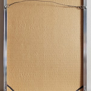 May include: Back of a framed artwork with a silver metal frame. The backing is a textured, light brown material. Black corner protectors and a wire for hanging are visible.