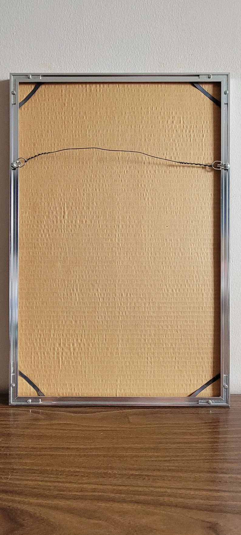 May include: Back of a framed artwork with a silver metal frame. The backing is a textured, light brown material. Black corner protectors and a wire for hanging are visible.
