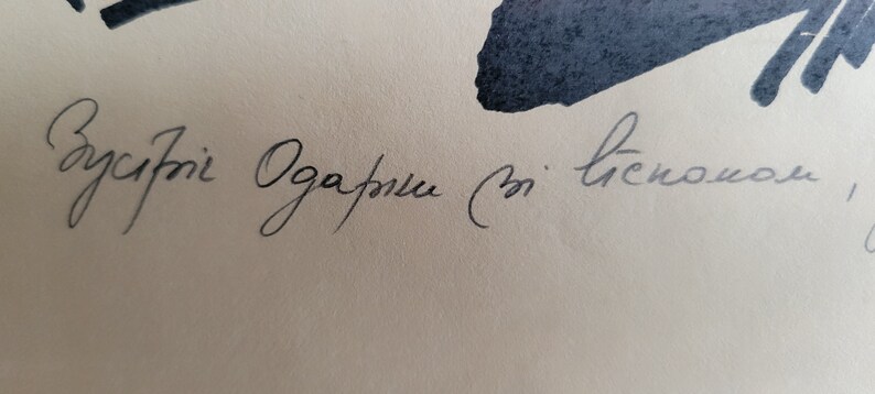 May include: Close-up of a handwritten script in black ink on a cream-colored paper. The script appears to be in a cursive style, with the text partially visible. The image also features dark blue ink blotches.