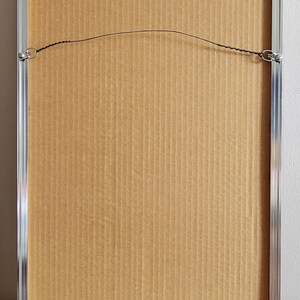 May include: Back of a framed artwork with a silver metal frame. The artwork is secured with a wire and metal hardware. The backing is made of corrugated cardboard.