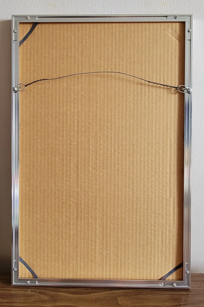 May include: Back of a framed artwork with a silver metal frame. The artwork is secured with a wire and metal hardware. The backing is made of corrugated cardboard.