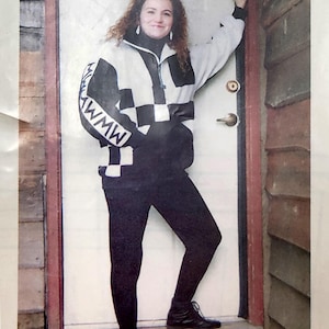 Puede incluir: Una mujer con una chaqueta a cuadros en blanco y negro con el texto "MWMW" en la manga. Está de pie frente a una puerta blanca con marco rojo. Lleva pantalones negros y botas negras.