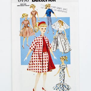 May include: A vintage Butterick sewing pattern with illustrations of doll outfits. The pattern shows various outfits, including dresses, a suit, and a bridal gown. The color scheme includes red, white, blue, and black.