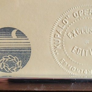 May include: A framed artwork with a silver metal frame. The artwork features two circular emblems and embossed text that reads "KUPALO OPERA PREMIERE EXCLUSIVE EDITION EDMONTON". The left emblem has a crescent moon and a laurel wreath.