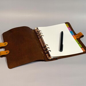 May include: A brown leather bound journal with a gold ring binder and a black pen on top of the open pages. The journal has a yellow leather strap with a button closure.