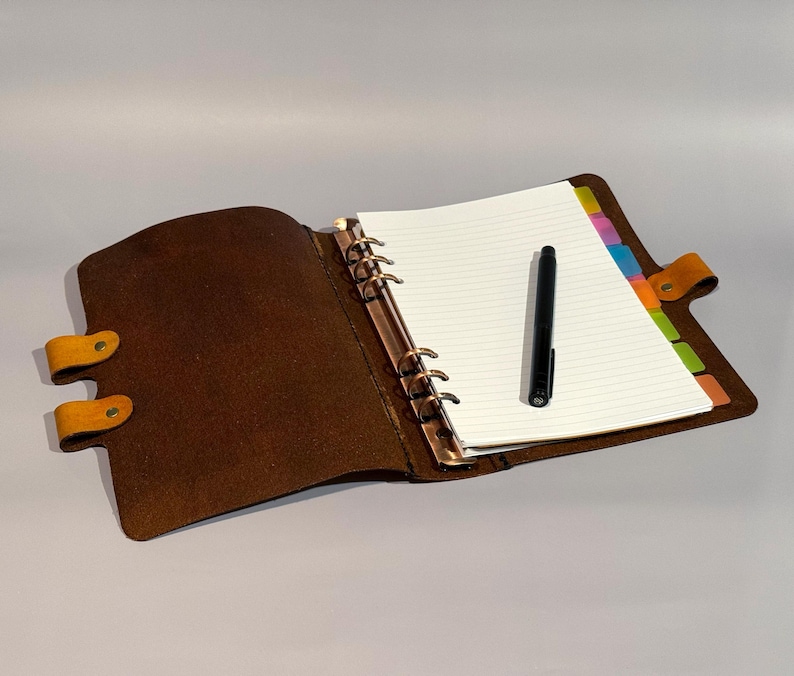 May include: A brown leather bound journal with a gold ring binder and a black pen on top of the open pages. The journal has a yellow leather strap with a button closure.