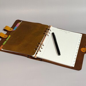 May include: A brown leather bound planner with a black pen and colourful tabs. The planner has a ring binding system and is open to a page with lined paper. The page has the text "To-Do for January 2023".