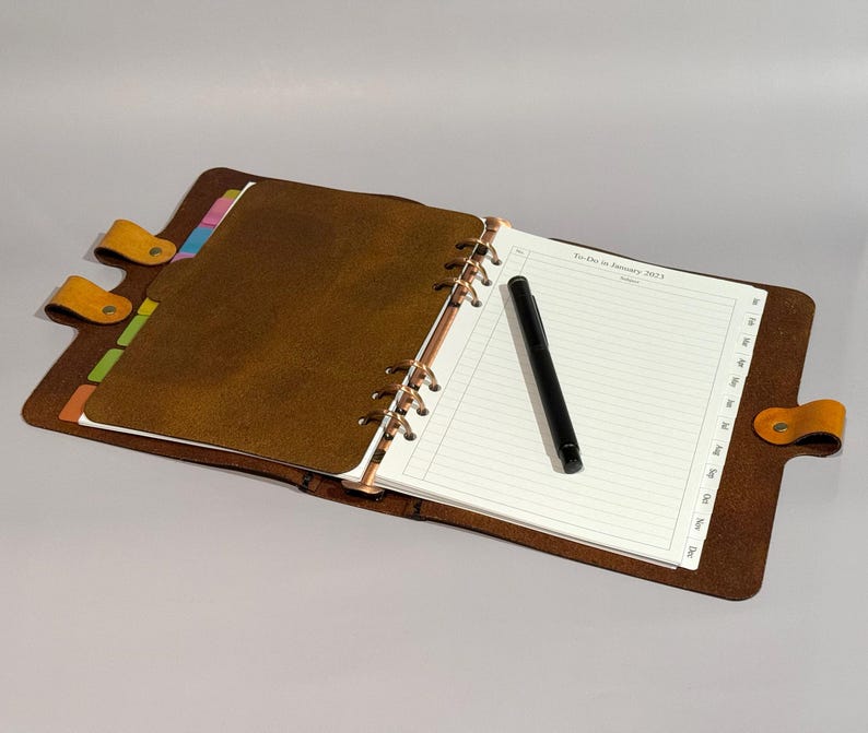 May include: A brown leather bound planner with a black pen and colourful tabs. The planner has a ring binding system and is open to a page with lined paper. The page has the text "To-Do for January 2023".