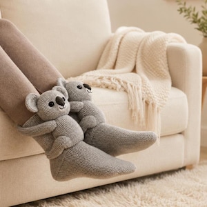 May include: A pair of light gray knit socks featuring plush koala bear figures attached to the ankles. The koalas have round ears, black noses, and embroidered eyes, with their arms wrapped around the front of the socks.
