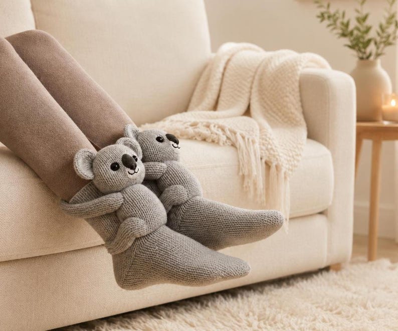 May include: A pair of light gray knit socks featuring plush koala bear figures attached to the ankles. The koalas have round ears, black noses, and embroidered eyes, with their arms wrapped around the front of the socks.