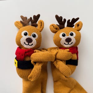 May include: Two matching knitted reindeer plush toys, each featuring a golden-brown body, dark brown antlers, a red scarf, and a black belt with a yellow buckle. The reindeer have white embroidered muzzles, dark brown noses, and black plastic eyes.