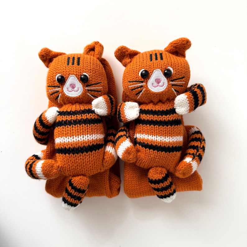 May include: A pair of orange knitted socks featuring a three-dimensional tiger design on the front. Each sock has a stuffed tiger head with black plastic eyes, embroidered whiskers, and a striped body with white and black accents on the paws.
