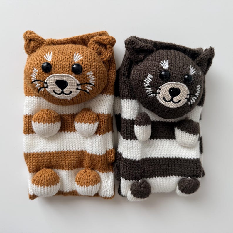 May include: A pair of knit socks featuring animal faces with black button eyes and embroidered details. One sock is brown and white striped, the other is dark brown and white striped. Both socks have small stuffed paws attached to the front.