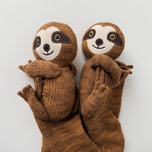 May include: Two brown knitted sloth toys with cream-colored faces, dark brown eye patches, and embroidered black features. Each sloth has a rounded head, long arms wrapped around its body, and a long, textured body extending downward.