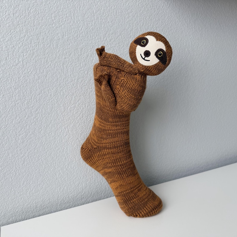 May include: A brown knitted sock featuring a sloth head attached to the cuff. The sloth has a cream-colored face with dark brown eye patches, a black nose, and a stitched mouth. The sock body is a variegated brown yarn with a ribbed texture.