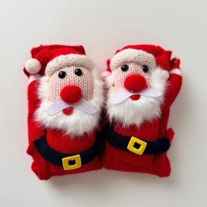 May include: A pair of red knit socks featuring a plush Santa Claus face on each. The Santa design includes a white knit hat, a fuzzy white beard, a red nose, black bead eyes, and a black belt with a yellow square buckle across the midsection.