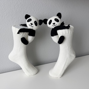May include: A pair of white knit socks, each featuring a small, plush panda bear attached to the cuff. The pandas are black and white with embroidered facial features, and their arms are positioned as if they are hugging the top of the socks.