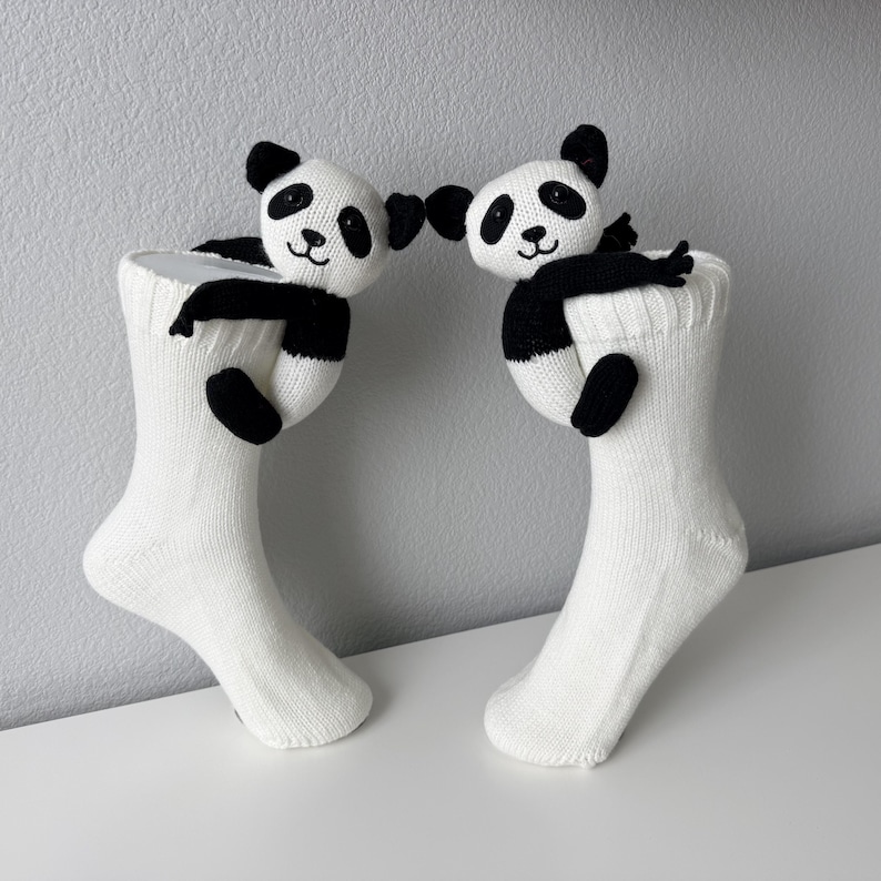 May include: A pair of white knit socks, each featuring a small, plush panda bear attached to the cuff. The pandas are black and white with embroidered facial features, and their arms are positioned as if they are hugging the top of the socks.