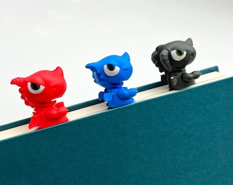May include: Three colorful dragon-shaped book markers. The markers are red, blue, and black, each with a unique expression and white and black eyes. They are placed on the edge of a teal book.