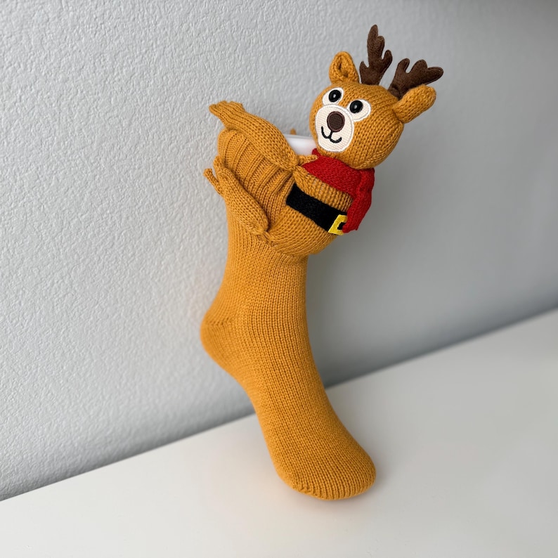 May include: A mustard yellow knit Christmas stocking featuring a plush reindeer character attached to the top. The reindeer has brown antlers, a red scarf, and a black belt with a yellow buckle. The stocking is made of a textured knit fabric.