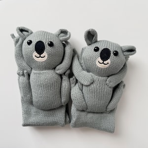 May include: Two gray knitted koala-shaped socks featuring embroidered black noses, black eyes, and light-colored muzzles. The koala faces are positioned at the top of each sock, with arms and legs extending down the length of the knit fabric.
