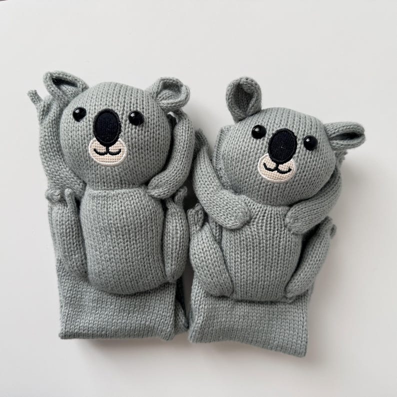 May include: Two gray knitted koala-shaped socks featuring embroidered black noses, black eyes, and light-colored muzzles. The koala faces are positioned at the top of each sock, with arms and legs extending down the length of the knit fabric.
