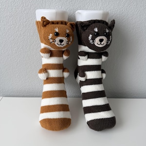 May include: A pair of knit socks featuring a three-dimensional red panda head and small paws at the top. One sock is brown and white striped, while the other is dark charcoal and white striped. Both socks have embroidered facial details.