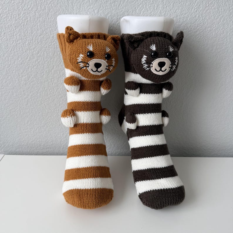 May include: A pair of knit socks featuring a three-dimensional red panda head and small paws at the top. One sock is brown and white striped, while the other is dark charcoal and white striped. Both socks have embroidered facial details.