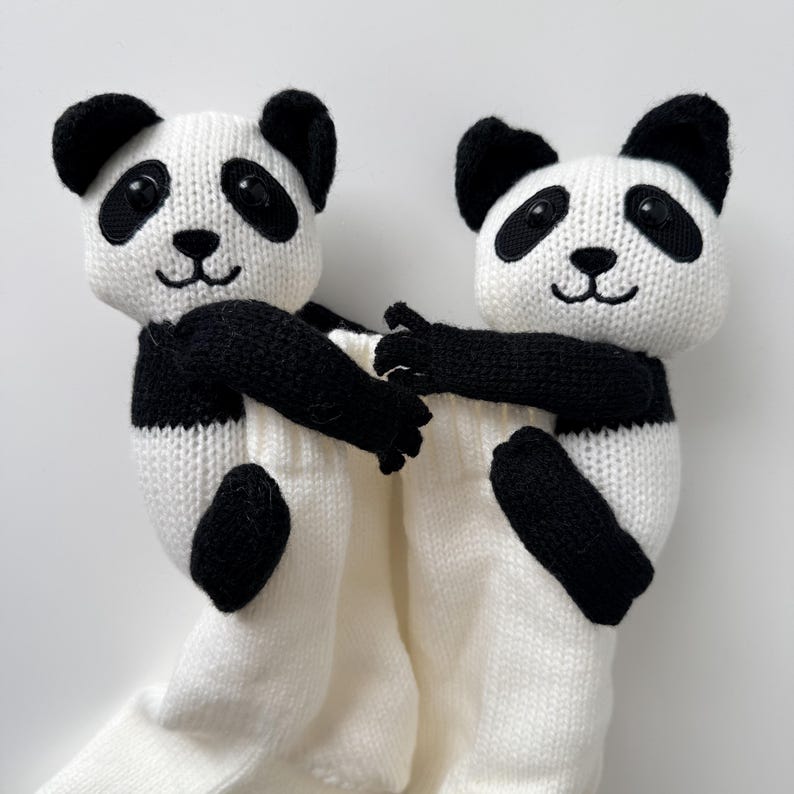 May include: Two knitted panda plush toys with black and white yarn, featuring round black eyes, embroidered noses, and mouths. The pandas are attached to the top of a pair of plain white knitted socks, with their arms reaching forward.