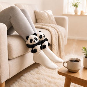 May include: A pair of white crew socks featuring plush panda bear attachments on the cuffs. The pandas are black and white with embroidered facial features and small arms that wrap around the front of the socks, worn over gray leggings.