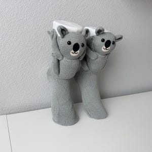 May include: A pair of light gray knit socks featuring three-dimensional koala bear designs on the cuffs. Each koala has embroidered black eyes, a black nose, and a small smiling mouth, with plush arms extending from the sides of the socks.